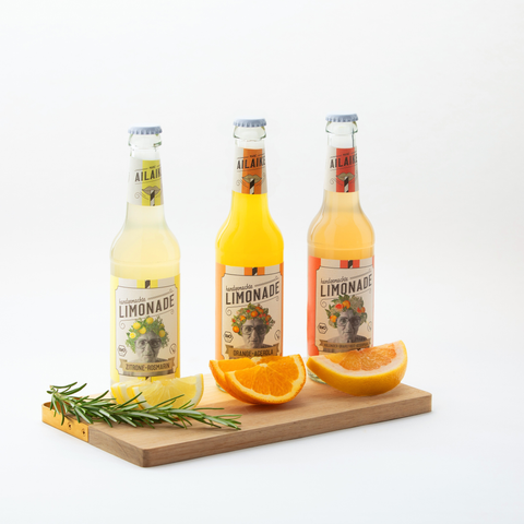 AiLaike Bio Limonade in Glasflaschen – Sorten Zitrone-Rosmarin, Orange-Acerola und Holunder-Grapefruit auf Holzbrett mit frischen Zitrusfrüchten und Rosmarin dekoriert.