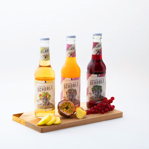 AiLaike Bio Schorle in Glasflaschen – Sorten Apfel, Maracuja-Ingwer und Schwarze Johannisbeere auf Holzbrett mit frischen Früchten dekoriert.