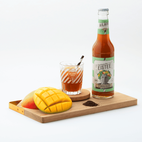 Bio-Eistee Pfirsich-Mango Flasche auf einem Brett mit einen gefüllten Glas und Pfirsich und Mango angerichtet.